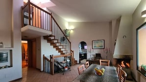 A spacious living area with a high ceiling, a staircase, a dining table with chairs, a fireplace, and a kitchen area.