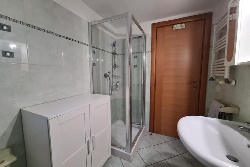 A bathroom with a glass shower enclosure, a wooden door, a white sink, and a cabinet.