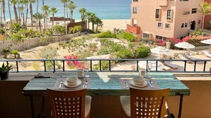 A balcony with two tables, one with a view of the beach and palm trees, and the other overlooking a landscaped garden with a building in the background. - Oceanview Penthouse Casa Del Mar (San José del Cabo)