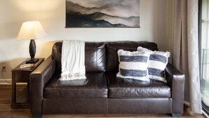 Een bruine leren bank met een witte plaid en gestreepte kussens, een lamp, en een muur met een berglandschap schilderij.