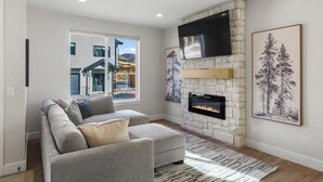 A modern living room with a stone fireplace, a large flat-screen TV, and a view of a building outside.