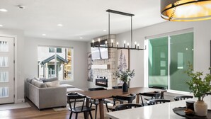 A modern living room with a sofa, dining table, and chairs. There is a fireplace, a TV mounted on the wall, and a large window.