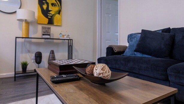 A modern living room with a dark blue sofa, a wooden coffee table with a remote control and decorative items, a wall-mounted lamp, and a framed artwork. - Modern 3-bed Bradley Stoke Self Check-in Free Parking Fast Wi-Fi (Bradley Stoke)