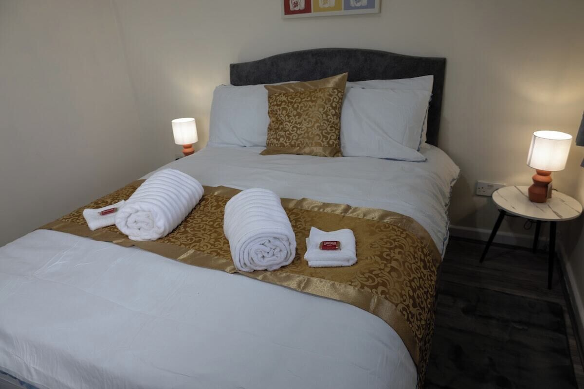 Ein ordentlich bezogenes Bett mit weißen Leinen und einer goldmusterten Tagesdecke, zwei Nachttischlampen und ein kleiner runder Tisch mit einer Lampe.