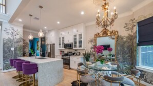 A modern kitchen with white cabinets, a central island, purple bar stools, and a dining area with a glass table and chairs.