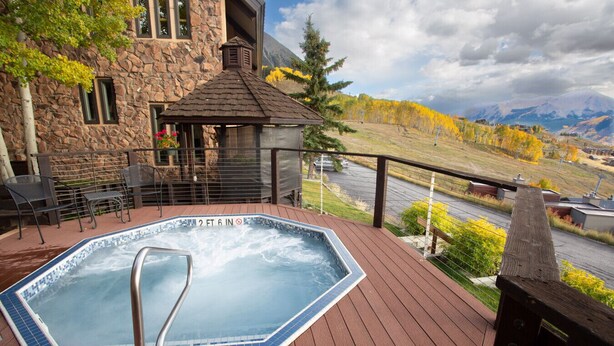 A hot tub on a wooden deck with a stone building in the background and a view of autumn trees and mountains. - 10 Steps to the Slopes! Unbeatable Ski-In/Ski-Out! Hot Tub! (Mount Crested Butte)