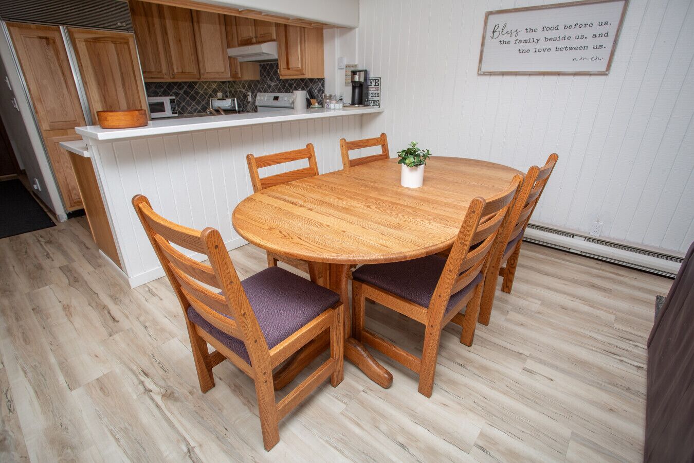 A dining area with a round wooden table and four chairs, a kitchen in the background, and a wall with a framed inspirational quote.