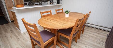 A dining area with a round wooden table and four chairs, a kitchen in the background, and a wall with a framed inspirational quote.
