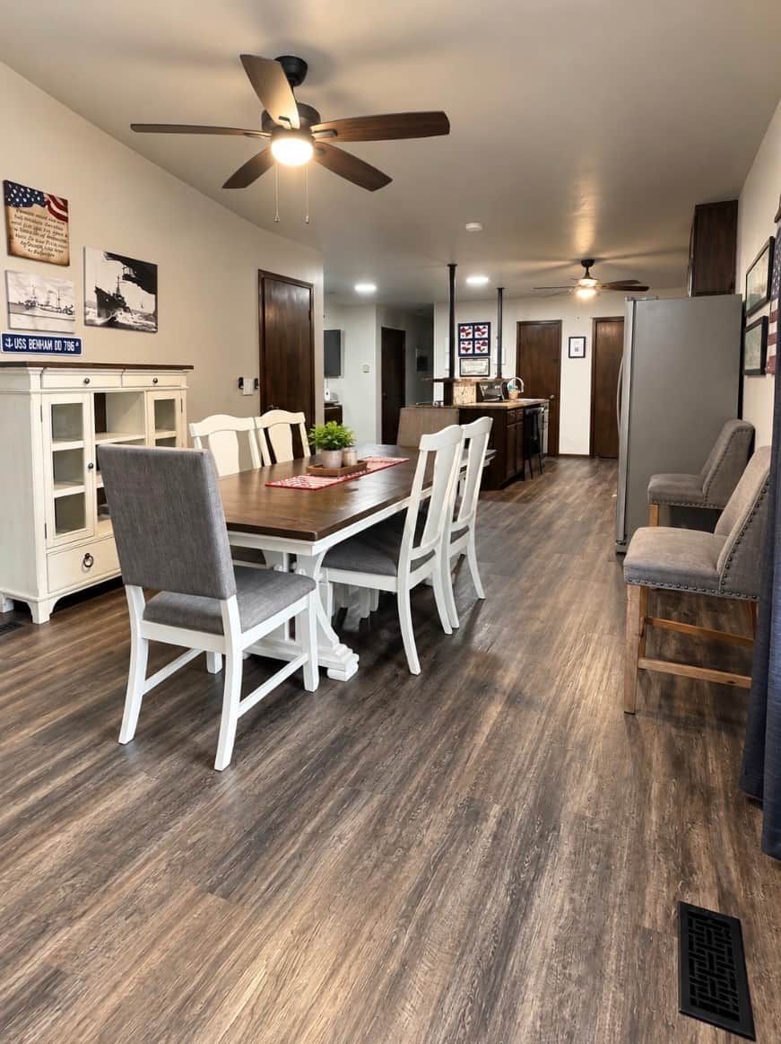 A dining area with a wooden table and chairs, a ceiling fan, and a kitchen in the background.