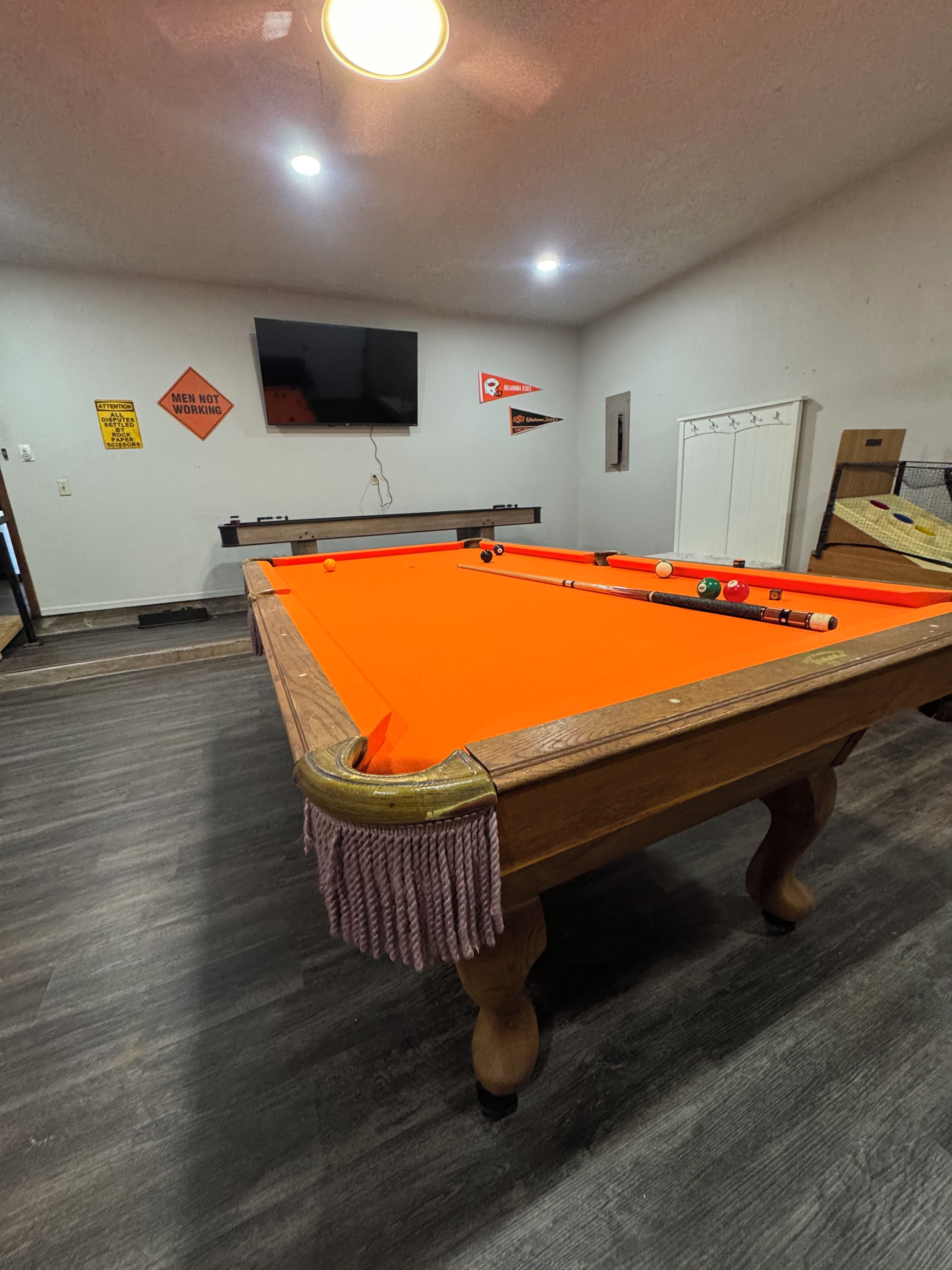 A pool table with a wooden frame and orange felt, a television mounted on the wall, and a door with a window.