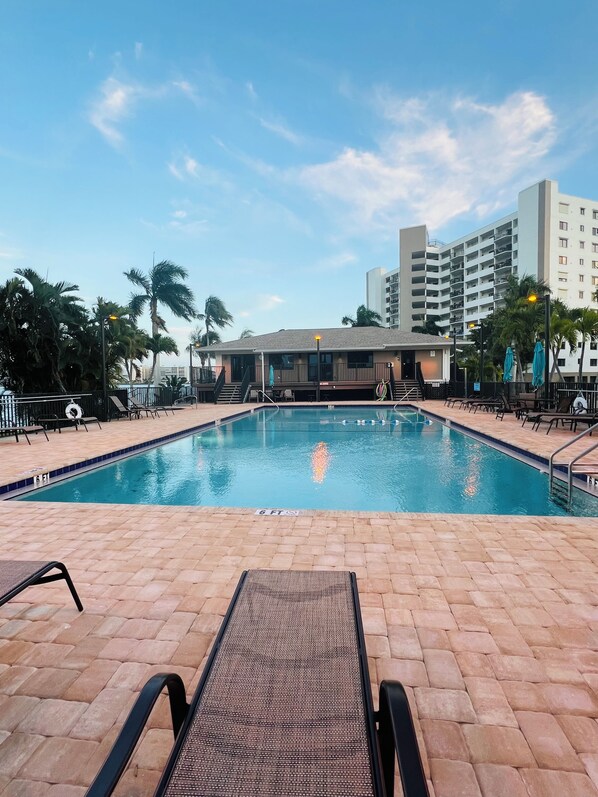 A swimming pool with lounge chairs, palm trees, and a building in the background.