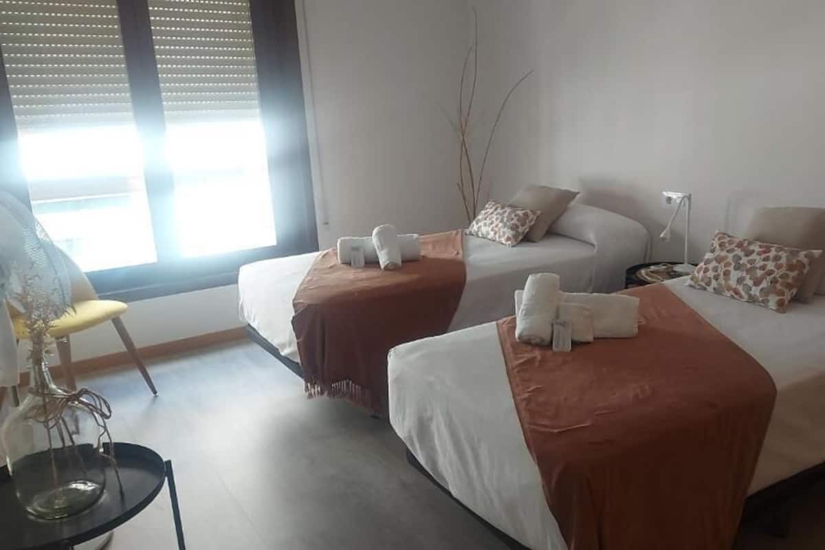 A hotel room with two beds, each with a brown blanket and white towels. There is a bedside table with a lamp and a chair with a fan.