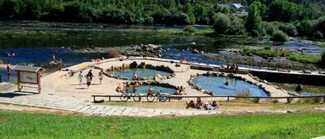 Un área de aguas termales naturales con varias piscinas rodeadas de construcciones de piedra, una plataforma de madera y un primer plano con césped.