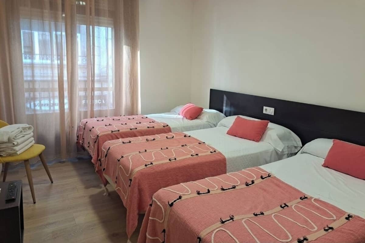 A hotel room with three beds, each with a pink and beige patterned bedspread, and a chair with folded towels.