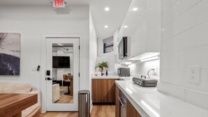 Una cocina moderna con encimeras blancas, armarios de madera y un cubo de basura de acero inoxidable, además de un letrero de salida visible.