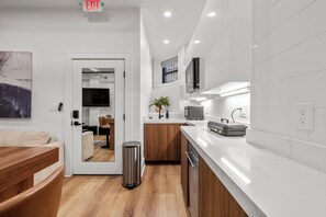 Una cocina moderna con encimeras blancas, armarios de madera y un cubo de basura de acero inoxidable, además de un letrero de salida visible.