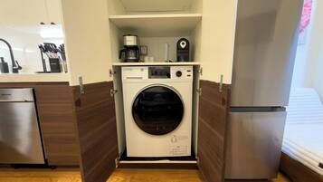 A compact laundry room with a front-loading washing machine, a coffee maker, and a small shelf with kitchen utensils.