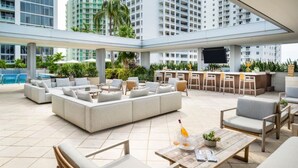 A spacious outdoor lounge area with a large sofa, dining tables, and chairs, surrounded by greenery and overlooking a swimming pool.