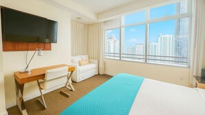 A hotel room with a bed, a desk, a chair, a TV, and a large window with a city view.