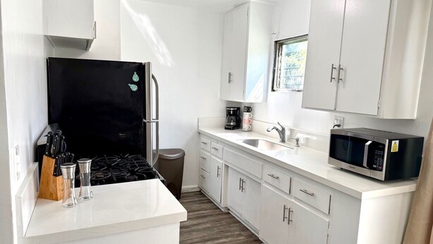 Une cuisine de taille réduite avec des meubles blancs, un réfrigérateur noir, un four à micro-ondes, une cuisinière à gaz et un évier.