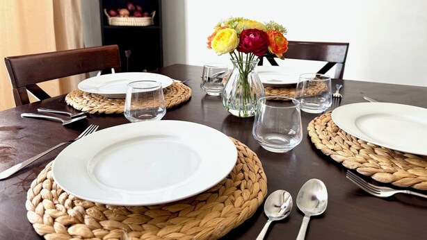 Een eettafel gedekt met witte borden, bestek en glazen, met geweven placemats en een vaas met bloemen.