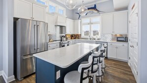 A modern kitchen with a large island, stainless steel appliances, and a blue accent wall. - Luxurious, Newly Furnished Home w/ stunning views & just steps from the beach (Ocean Isle Beach)