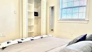 Et soveværelse med seng, walk-in closet, et vindue med persienner og et lille spejl.