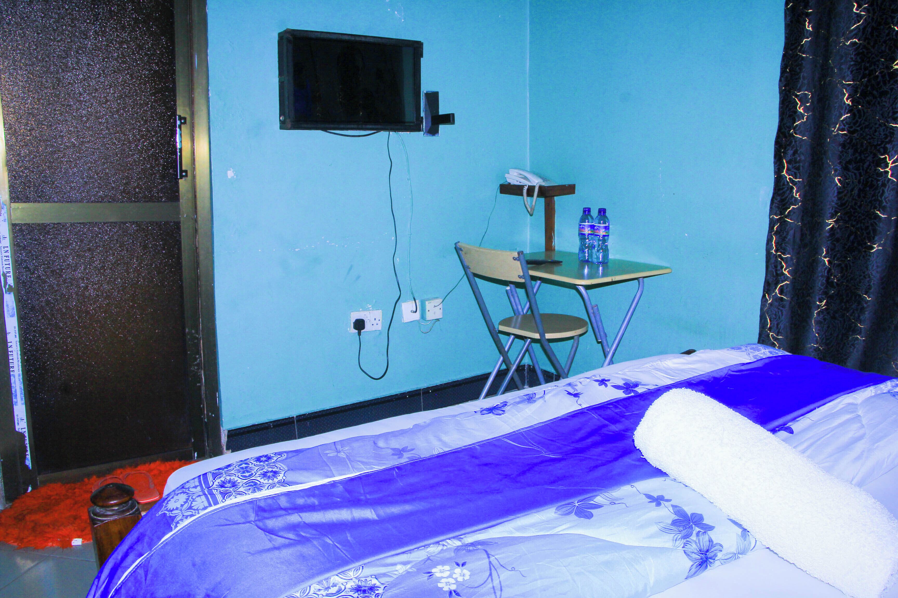 Una habitación con una cama, una silla, una mesita pequeña con un teléfono y botellas de agua, y un televisor colgado en la pared.
