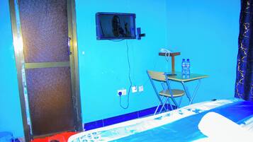 Una habitación con cama, un escritorio con silla, un televisor en la pared y una botella de agua sobre el escritorio.
