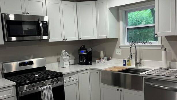 A modern kitchen with white cabinets, stainless steel appliances, a marble countertop, and a window with a view of greenery.