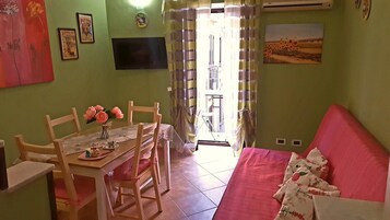 Un comedor con mesa y sillas, un televisor montado en la pared y un sofá con cojines estampados.