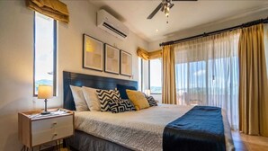 A bedroom with a bed, bedside table, lamp, air conditioning unit, ceiling fan, and large windows with curtains. - Gorgeous Ocean View Rooftop Patio, Private gated 2 bedroom home, Walk to beach (Coco)