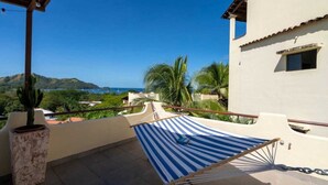 A balcony with a striped mat, a potted cactus, and a view of the sea and hills. - Gorgeous Ocean View Rooftop Patio, Private gated 2 bedroom home, Walk to beach (Coco)