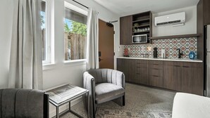 Un espacio habitable compacto con kitchenette, un sillón gris, una mesita y vistas al exterior a través de una ventana.