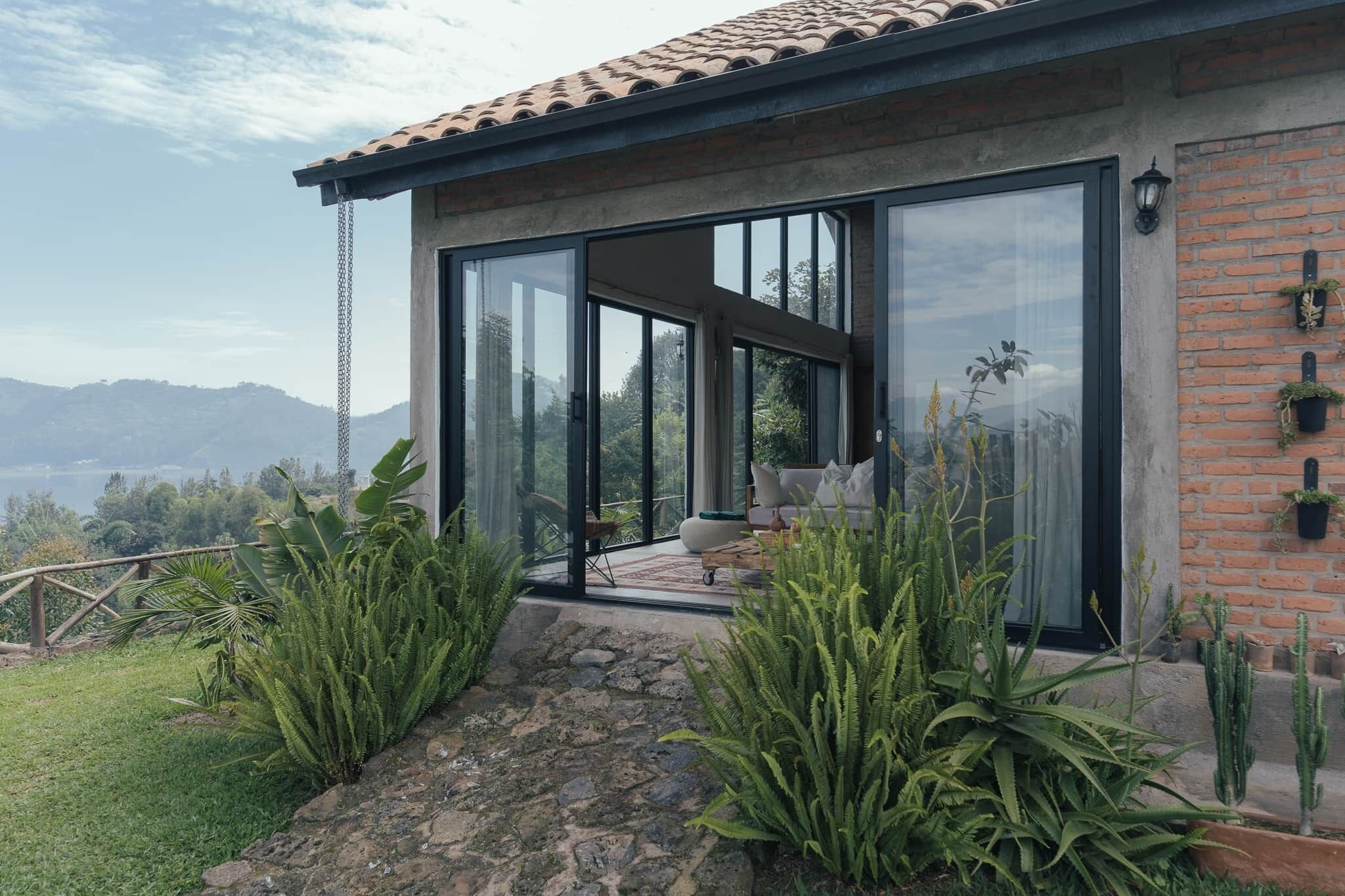 A modern house with a glass door leading to a balcony, surrounded by lush greenery and a mountainous backdrop.