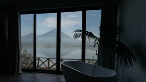 A bathroom with a freestanding bathtub, a large window with a view of a lake and mountains, and a potted plant. - The Writer's Patch - Two bedroom house on lake  Ruhondo (Musanze)