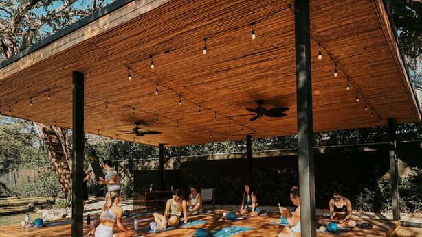Ein Yoga-Kurs im Freien unter einer Holzhütte mit Deckenventilator, umgeben von viel Grün.