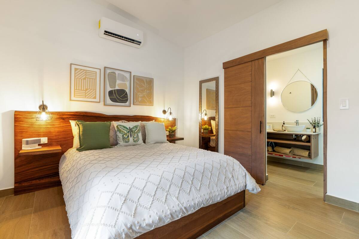 Un dormitorio con una cama grande, cabecera de madera, mesitas de noche, una obra de arte en la pared y un baño visible a través de una puerta abierta.