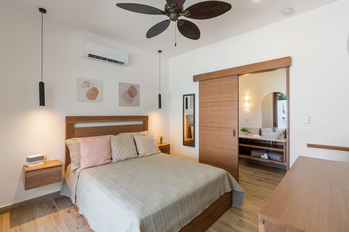 Un dormitorio con cama, mesitas de noche, ventilador de techo, aire acondicionado instalado en la pared, obras de arte y un baño visible a través de una puerta abierta.
