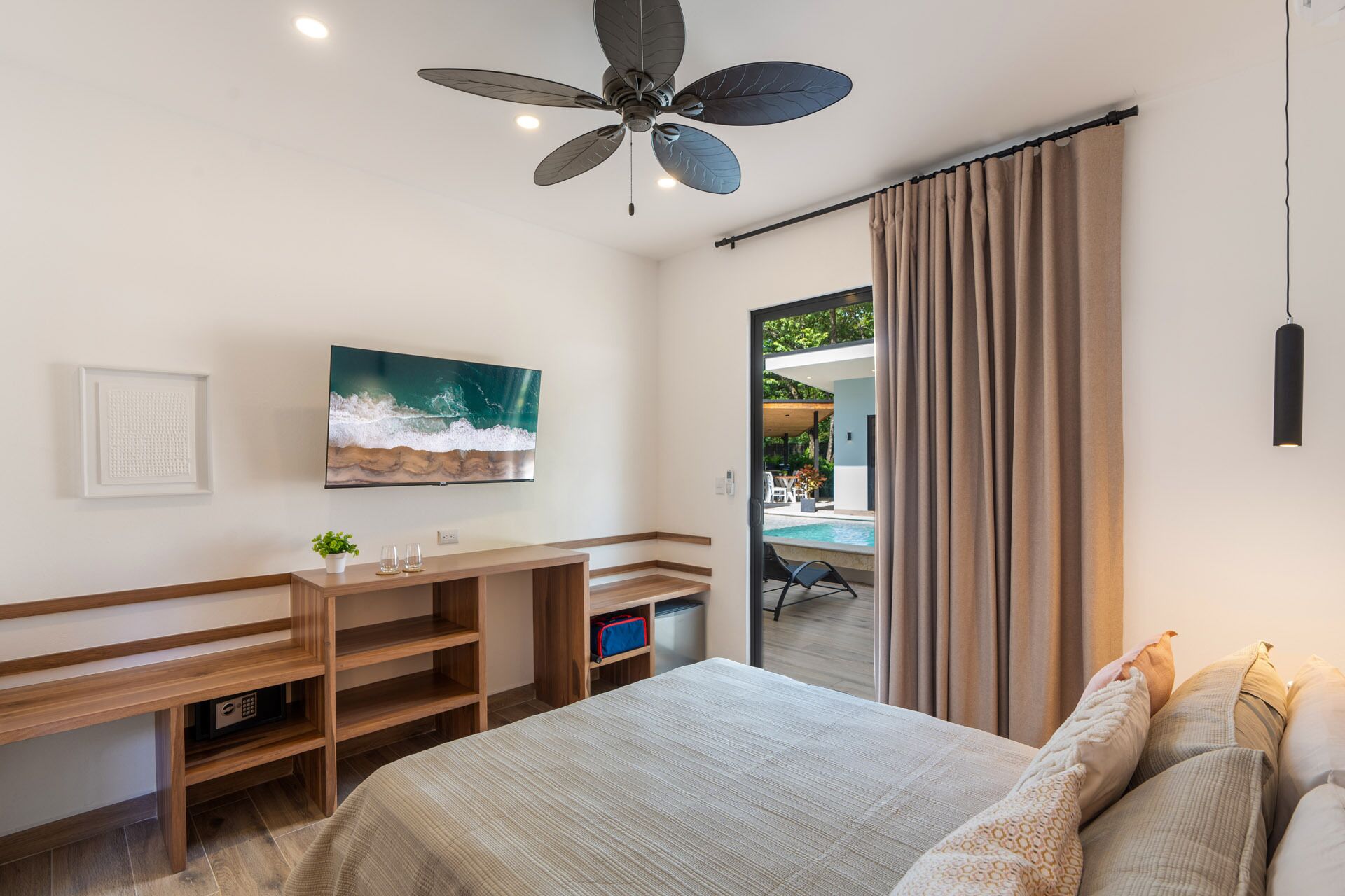 A modern bedroom with a bed, a desk, a television, and a view of an outdoor pool area.