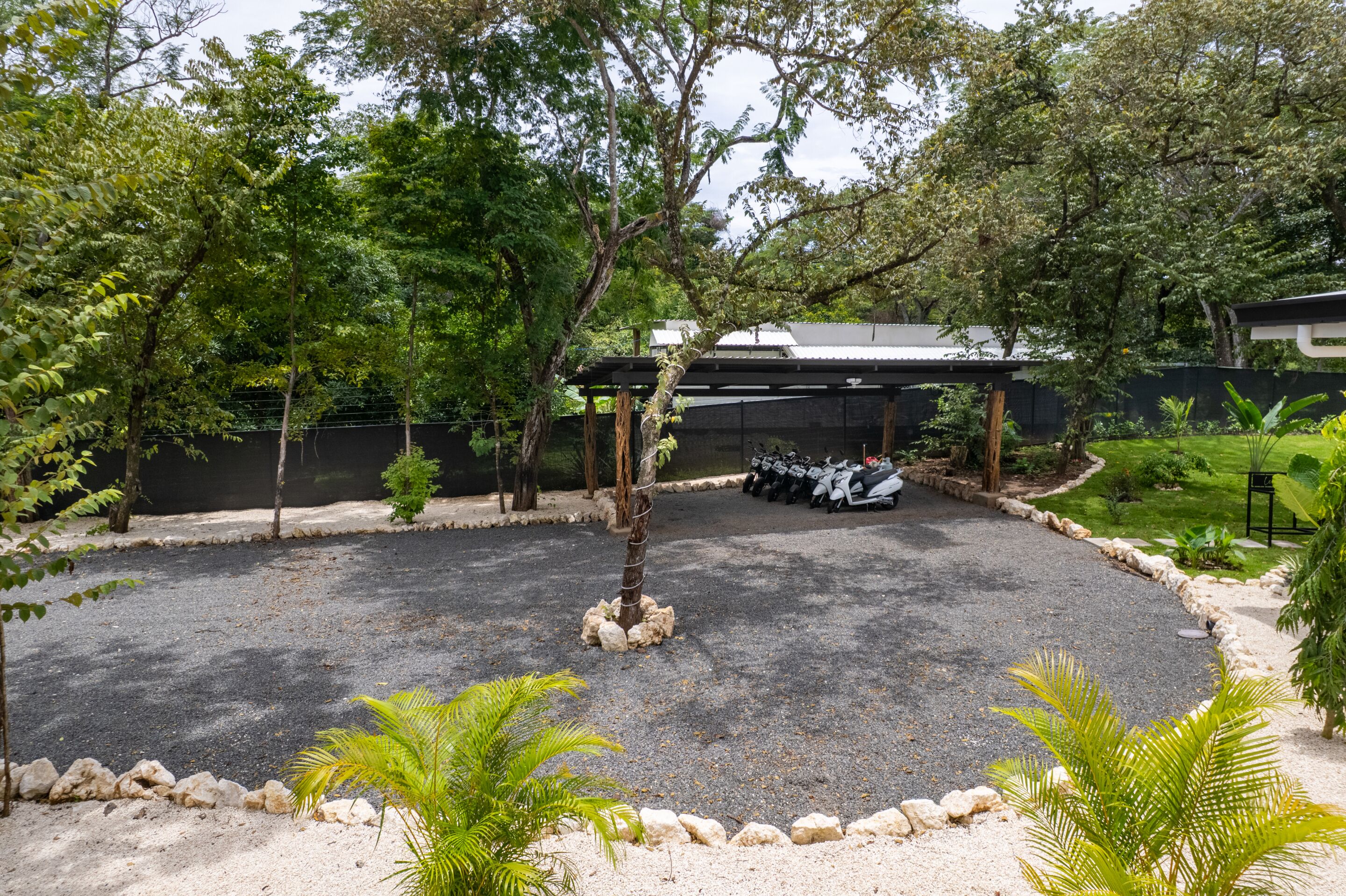 A gravel parking area with a row of parked scooters, surrounded by trees and a building with a metal roof.