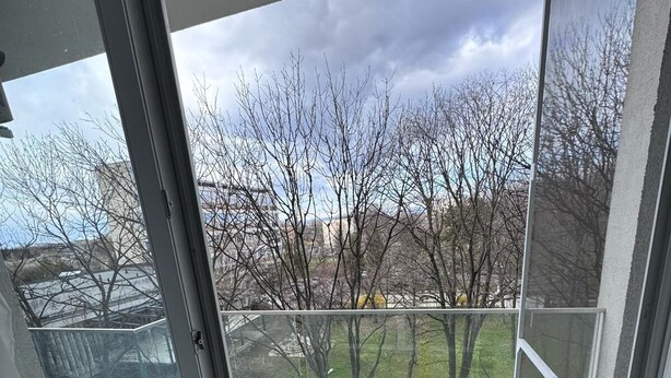 Vista da un balcone con porta vetrata, su una zona verde con alberi e edifici sullo sfondo.