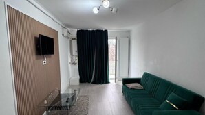 Un soggiorno moderno con un divano verde, una TV a schermo piatto montata su un pannello di legno, un tavolino in vetro e una ventola a soffitto.