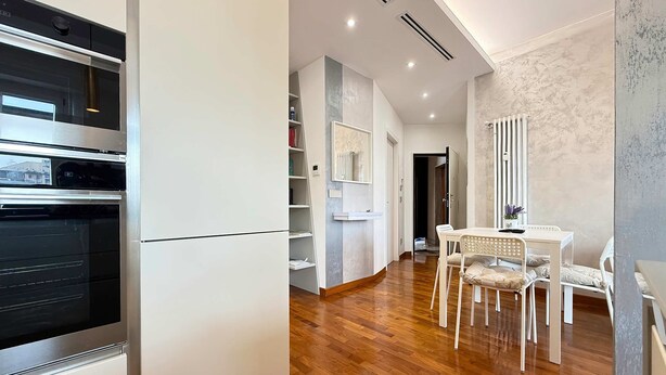 A modern kitchen with a built-in oven, a dining area with a white table and chairs, and a hallway with a radiator and shelves. - Casa Laura in Milano (Milan)
