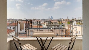 Un balcone con tavolo e sedie in legno, vista su una città con edifici e cielo sereno.