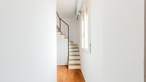 Un corridoio stretto con pavimento in legno e pareti bianche, che conduce a una scala con gradini in legno e ringhiera nera.