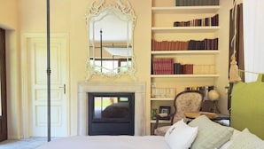 A bedroom with a large bed, a fireplace, a bookshelf, and a mirror.