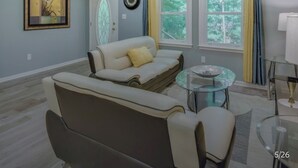 Una sala de estar con un sofá, una mesa de centro de vidrio, un ventilador de techo y una ventana con cortinas.