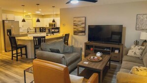 Ein modernes Wohnzimmer mit einer Couch, einem Couchtisch und einem Fernseher. Es gibt eine Küchenzeile mit Schränken und einem Kühlschrank.