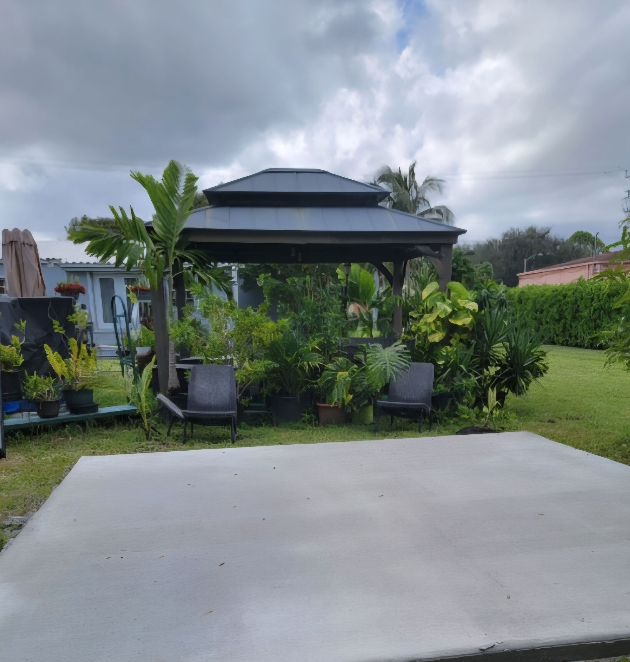Un espacio al aire libre con techo, con una mesa de hormigón, rodeado de plantas en macetas y sillas.